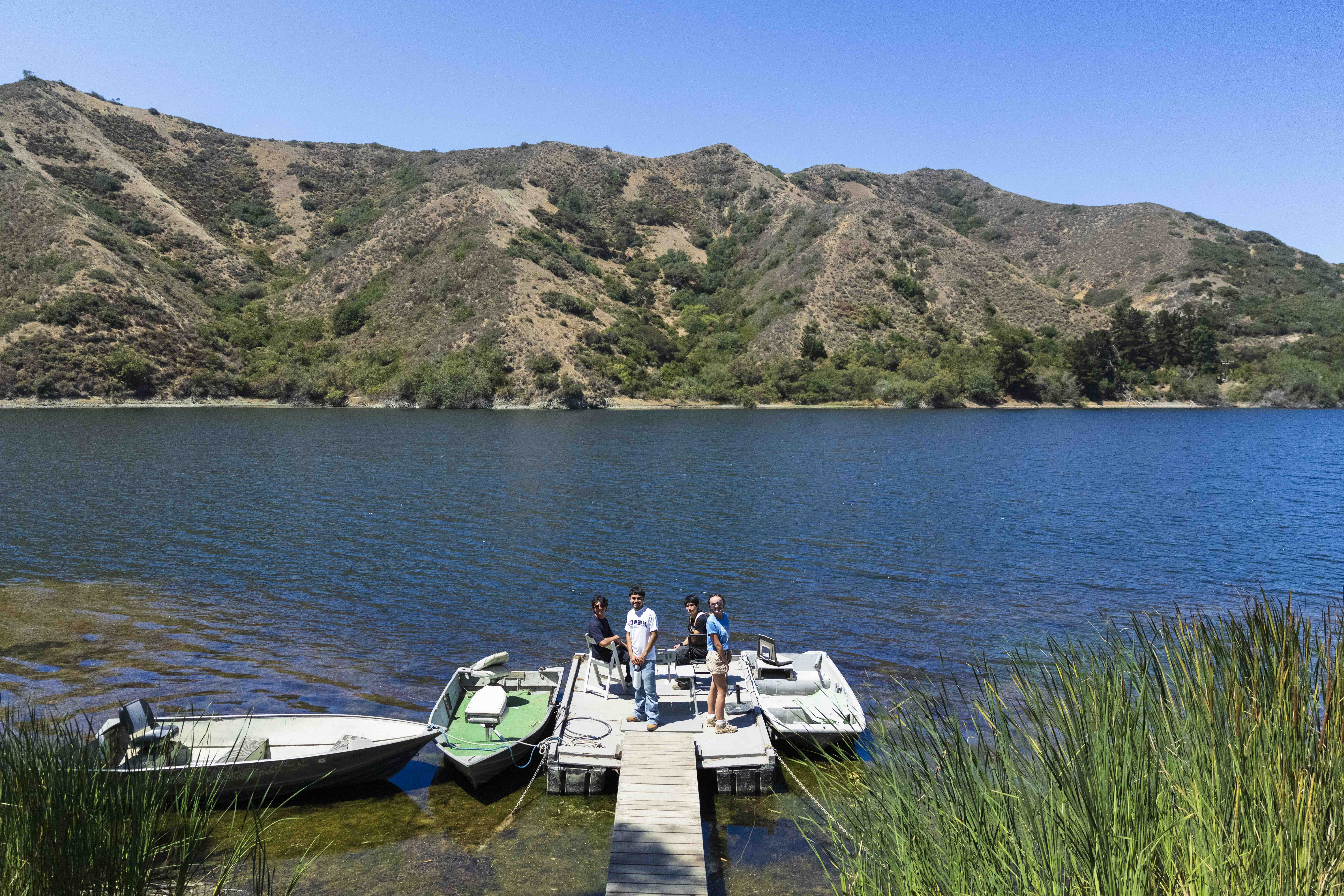 Four people stand on a small floating dock with boats moored on a mountain-ringed reservoir
